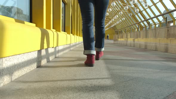 Women's Legs in Burgundy Boots and Blue Jeans Walk on a Closed Pedestrian Bridge alt
