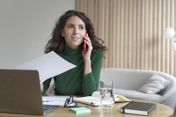 Smiling Italian businesswoman sitting at table making call at her workplace while working on laptop - Stock Photo - Images
