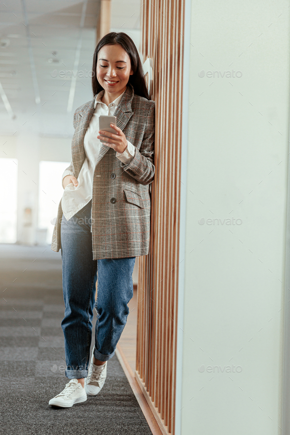 Portrait of smiling asian business woman standing in modern office with phone - Stock Photo - Images