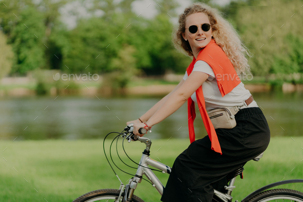 curly woman looks aside, rides sport bicycle, covers distance and travels in open air