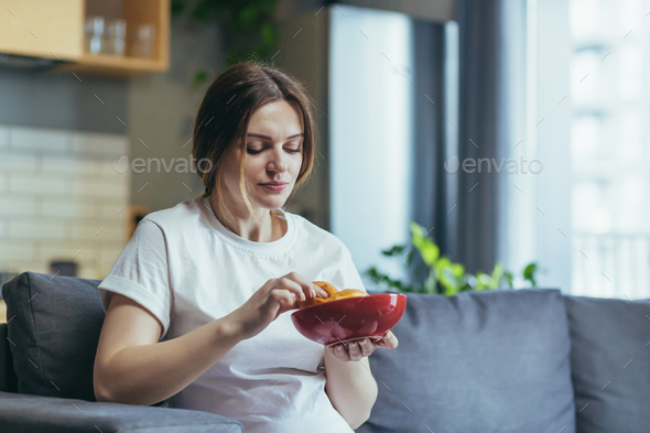 Pregnant woman at home sitting on sofa eating unhealthy and junk food donuts