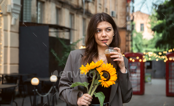 A young woman with a bouquet of sunflowers and a cold coffee drink in the city.