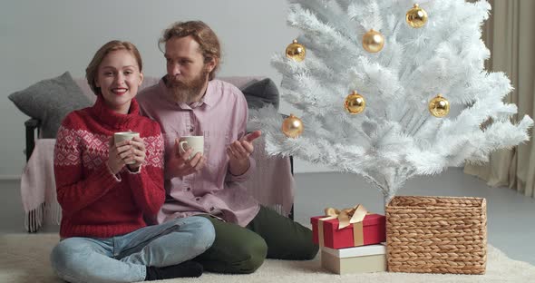 Young Couple Drinking Cocoa Cappuccino Tea While Sitting at Cozy Home on Floor Near Christmas Tree alt