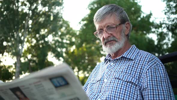Senior in Plaid Shirt Sits on a Bench in the Park and Reads a Newspaper alt
