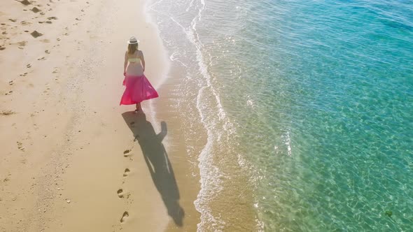 Happy Woman in Pink Beach Dress Walking By Scenic Tropical Ocean Beach at Sunset alt