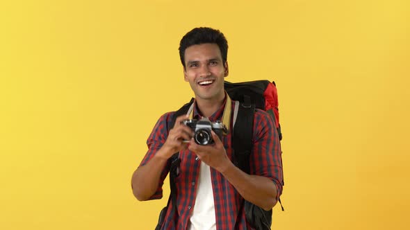 Young happy smiling Indian man with backpack and camera taking photos and giving thumbs up