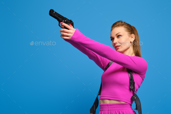 Stylish woman in purple knitted pantsuit aims a gun over blue studio background