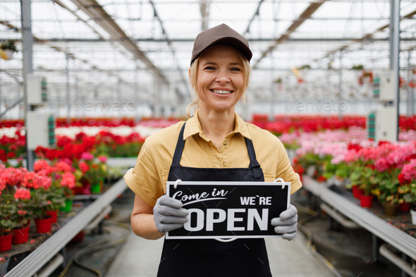 Pretty woman owner of a flower greenhouse with a sign come in we\'re open