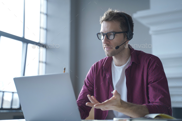 European man online tutor wearing glasses sitting at desk in headset and talking by video call