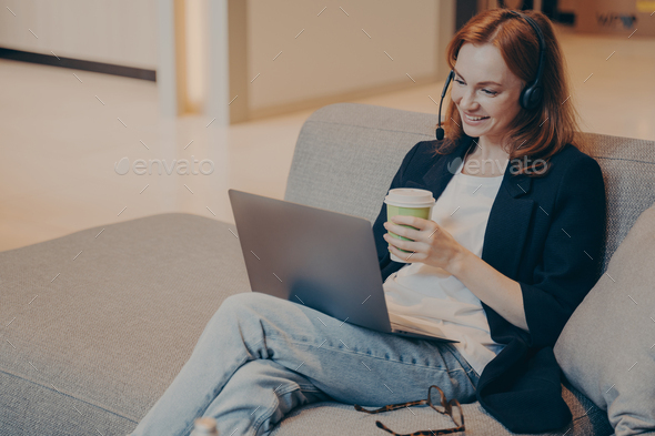 Smiling young female call center agent in wireless headset and laptop sitting on sofa in cafe