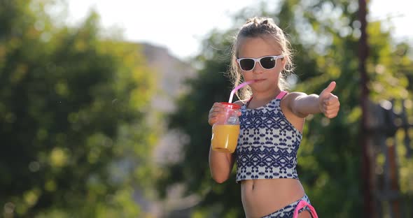 Beautiful little girl, cute toddler with sunglasses is drinking orange juice and shows thumb up. 