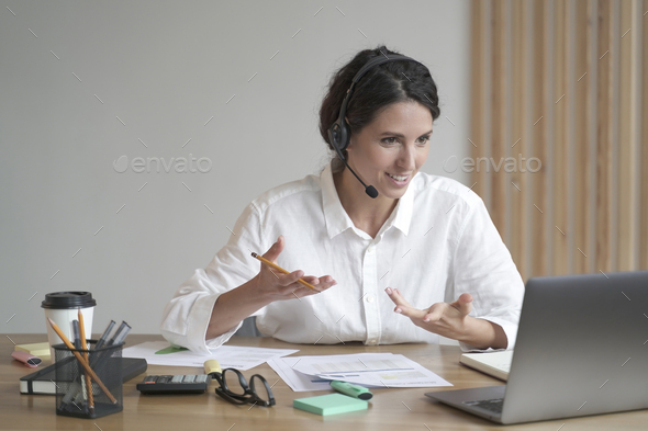 Smiling woman online tutor sitting at desk in headset and talking by video call on laptop computer