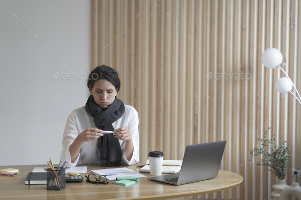 Sick young hispanic woman in scarf around neck looking at electronic thermometer at workplace