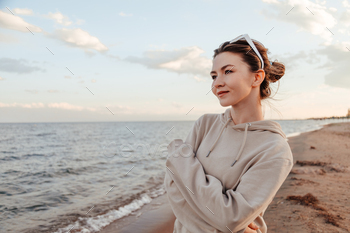 Woman standing by the lake shore in hoodie and sunglasses alone looking at waves