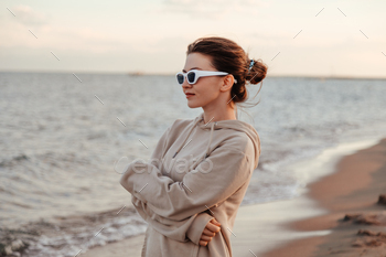 Woman standing by the lake shore in hoodie and sunglasses alone looking at waves