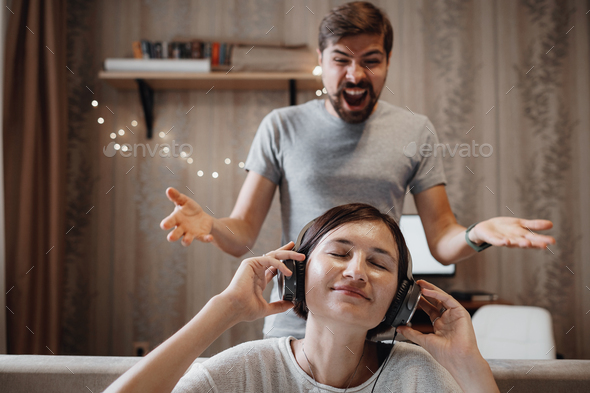 angry husband shouting at wife sitting on sofa and covering ears with headphones