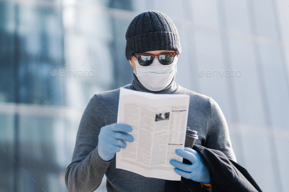 Shot of young man reads article about coronavirus symptoms, wears protective medical mask