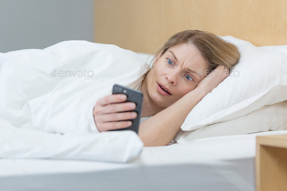 Woman lying in bed in the morning, shocked reading bad news from the phone