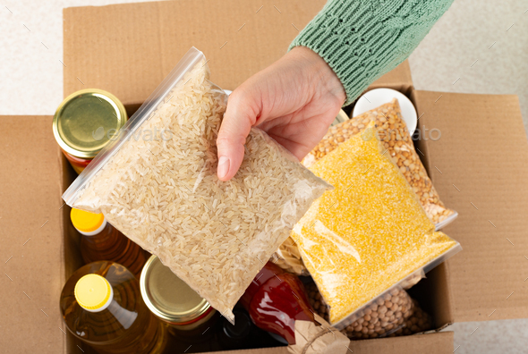 Plastic container with uncooked rice in female hand on emergency food box background