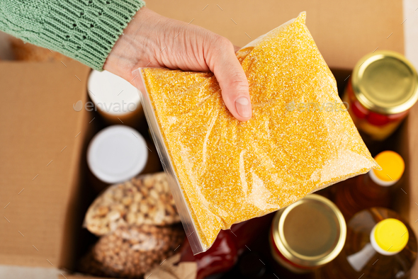 Plastic container with corn grits in female hand on emergency food box background