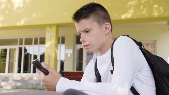 A Caucasian Teenage Boy Looks at a Smartphone and Acts Frustrated As He Sits in Front of School