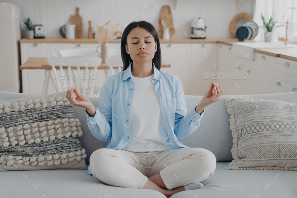 Calm woman meditate, practice yoga, sitting on sofa in lotus position ...