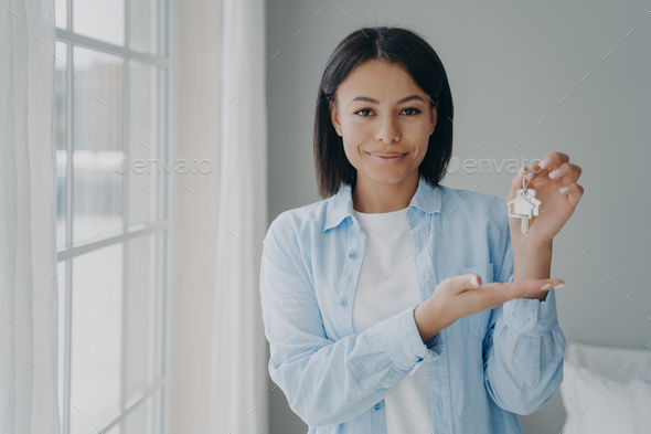 Satisfied hispanic girl holding house keychain in hand. Young woman is buying apartment and moving.