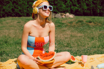 Stylish woman in headphones eats watermelon in outdoor in summer