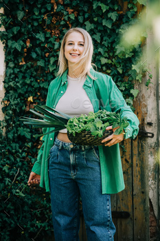 Stylish woman in green shirt with basket full of vegetables stands on ivy background