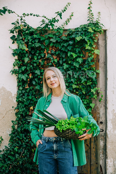 Stylish woman in green shirt with basket full of vegetables stands on ivy background