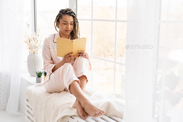 Smart intelligent clever african-american young woman reading a book in pink pajamas