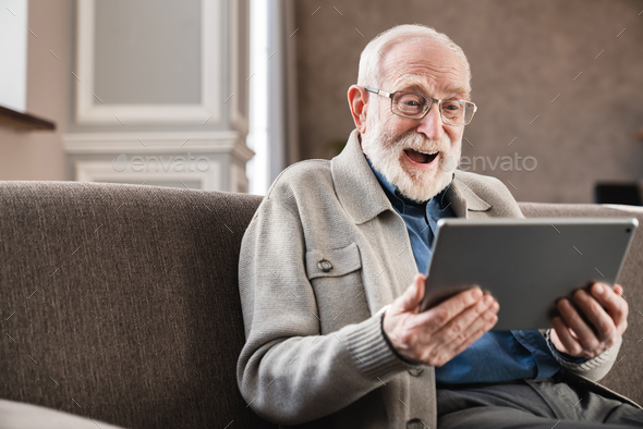 Laughing senior old grandfather using digital tablet on the sofa . Modern elderly pensioner