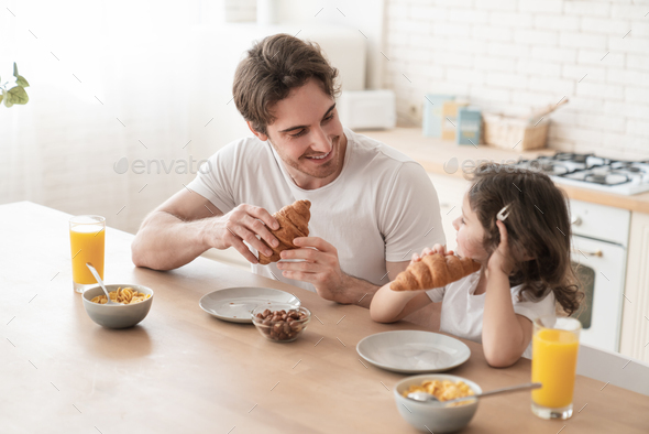 Caring young caucasian father dad eating croissants for breakfast together in the kitchen.