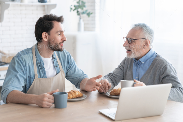 Communicating talking old elderly senior father with his adult son having breakfast using laptop