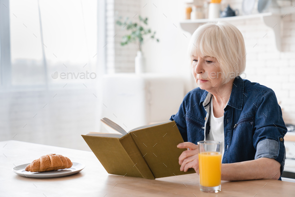 Aged elderly senior grandmother woman reading a book having breakfast in the kitchen.