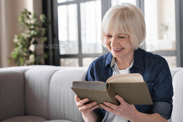Happy old aged elderly senior woman sitting on a sofa and reading an interesting book
