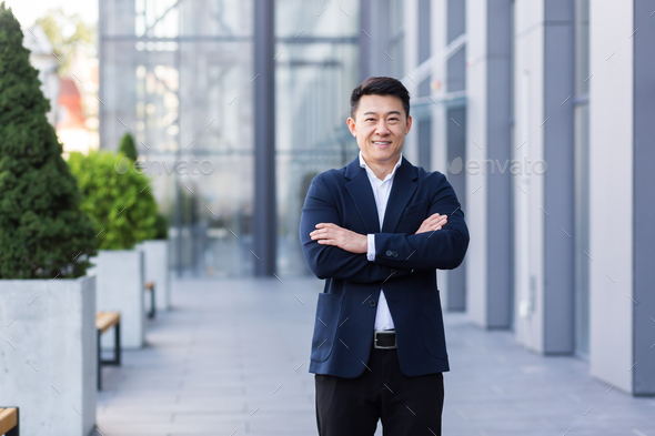 Successful Asian boss near the office looks at the camera with crossed arms smiles