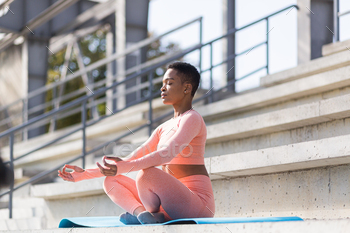 African American woman performs breathing exercises sitting on a yoga mat in the park