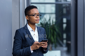 thinking business woman using phone, african american woman in business suit near office