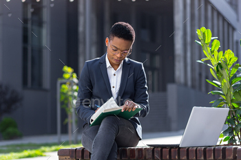Young beautiful female student studying, remotely in the park, sitting on a bench