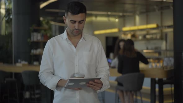 Portrait of Handsome Young Middle Eastern Man Using Tablet, Drinking Coffee, and Smiling at Camera alt