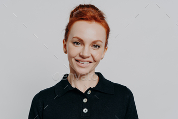 Smiling young woman with red hair in bun looking at camera isolated over grey background
