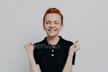 Happy euphoric red haired female with closed eyes holding clenched fists up and celebrating win