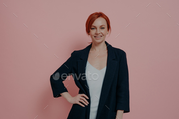 Confident beautiful redhead woman in navy jacket smiling at camera and keeping on hand on hip