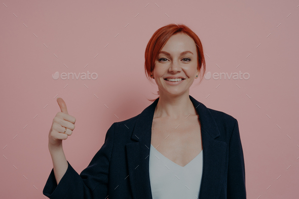 Young successful cheerful red-haired business woman showing thumb up hand gesture - Stock Photo - Images