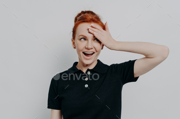 Happy young redhead woman with opened mouth touching her forehead with hand