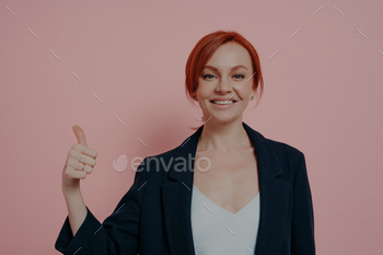 Young successful cheerful red-haired business woman showing thumb up hand gesture