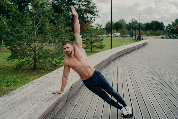 Athletic man stands in side plank pose, raises one arm, poses with shirtless torso