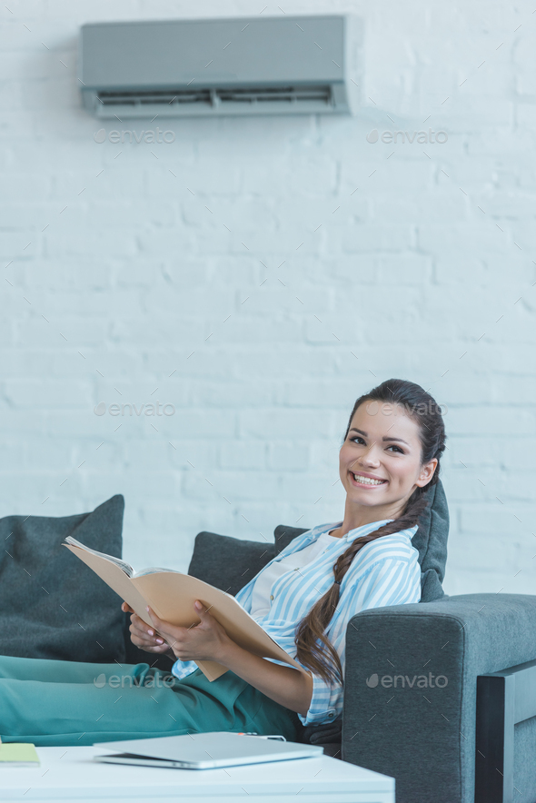 smiling woman with book sitting on sofa, with air conditioner on wall