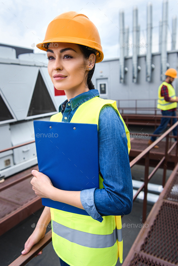 female architect in helmet holding clipboard on roof, male colleague
